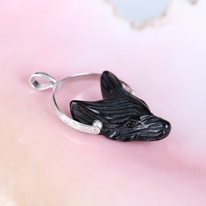 May include: Black onyx wolf pendant with a silver bail and a detailed carved design. The pendant is on a pink background.