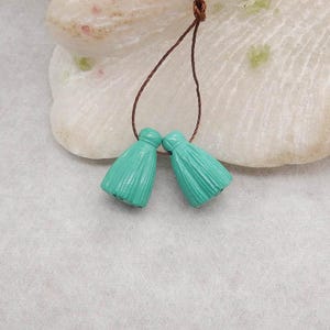May include: Two turquoise tassel beads hanging from a brown string. The tassels are ribbed and have a rounded top. The background is a light-coloured, textured surface.