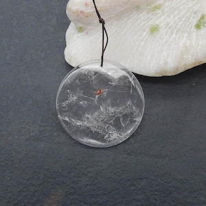 Peut inclure: Un pendentif rond et transparent avec des veines blanches internes et une petite inclusion rougeâtre. Le pendentif est suspendu par un cordon fin et brun foncé. Le fond comprend une pierre blanche texturée et une surface gris foncé.