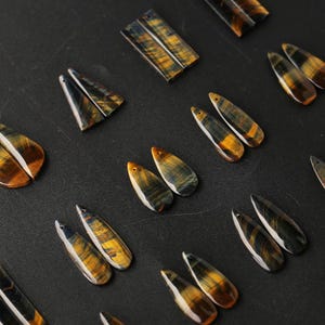May include: A collection of polished tiger's eye gemstone pendants in various shapes, including teardrops and rectangles. Each piece displays a chatoyant luster with alternating bands of golden-brown and dark blue, featuring small drilled holes for hanging.