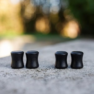 May include: Four black wooden ear plugs are arranged in a row on a concrete surface. The plugs have a cylindrical shape with a slightly flared middle section. The background is blurred, with hints of green and yellow.