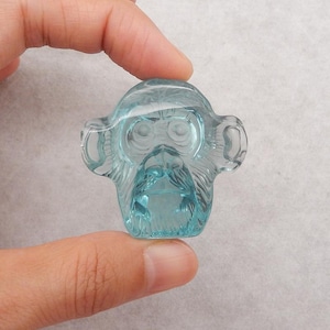 May include: A light blue, transparent monkey figurine. The monkey's face is intricately carved with detailed features, including eyes, nose, and mouth. The figurine is held between fingers, showcasing its size and clarity. The overall design is smooth and polished.