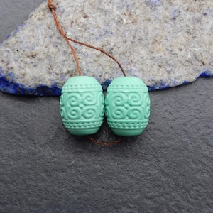 May include: Two pale green, oval beads with a detailed, swirling pattern. The beads are strung on a brown cord, set against a grey and blue stone background. These decorative beads are suitable for jewellery making.