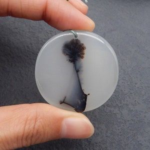 May include: A round, white pendant with a dendritic agate design. The agate features a dark brown and black tree-like pattern. A green cord is attached for wearing as a necklace. The pendant is held by a hand.
