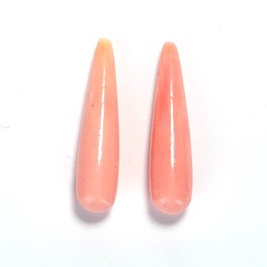 May include: Two polished, teardrop-shaped gemstones in a soft peach color. The gemstones are elongated with a rounded base and a tapered top. The surface of the stones has a glossy finish, reflecting light. The background is white.