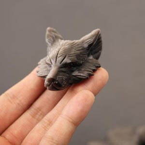 Pode incluir: Uma figura de cabeça de lobo esculpida à mão em pedra cinzenta escura com um acabamento liso e polido. A escultura apresenta texturas de pelo detalhadas, orelhas pontiagudas e pequenos olhos incrustados de cor vermelho-escura.