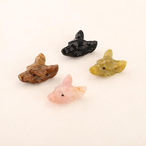 May include: Four carved stone animal head figurines in brown, black, pink, and yellow-green. Each head has detailed features and small holes, possibly for stringing. The figurines are arranged on a white surface.