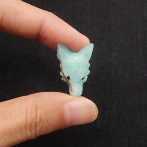 Puede incluir: Una pequeña figura tallada con forma de cabeza de lobo en color verde azulado claro. Los rasgos del lobo son detallados, con orejas puntiagudas y un hocico definido. La figura se sostiene entre dos dedos sobre un fondo oscuro.