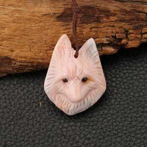 May include: A carved pendant in the shape of a fox's head, featuring a light pink and white striped pattern. The fox has dark brown eyes and a brown cord for hanging. The pendant is made of stone and is displayed against a dark background.