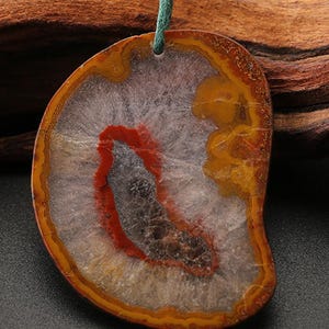 May include: A pendant made from a polished agate slice featuring concentric bands of amber, yellow, and white surrounding a central core of deep red and dark gray. The stone is suspended from a thin green cord threaded through a drilled hole.