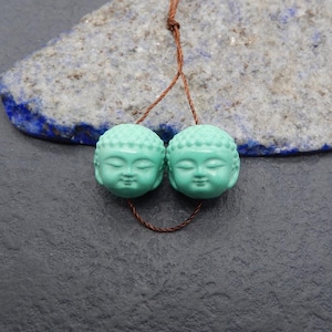 May include: Two turquoise Buddha head beads strung on a brown cord. The beads have detailed facial features and a serene expression. The background features a grey surface and a piece of blue and grey stone.
