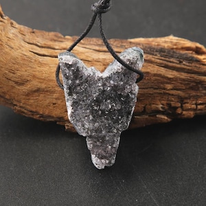 May include: A dark purple, crystalline druzy pendant shaped like a fox head, suspended from a thin black cord. The stone features a rough, textured surface with small, sparkling mineral points throughout, creating a distinct, angular silhouette.
