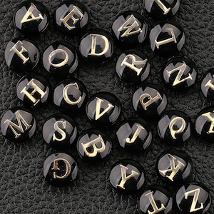 Pode incluir: Uma coleção de contas pretas e redondas com letras maiúsculas douradas. As letras estão em uma fonte serif e são centralizadas em cada conta. As contas estão dispostas em uma superfície escura e texturizada.