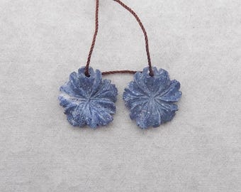 May include: Two blue flower-shaped pendants with intricate carved details, connected by a brown string. The pendants are a dusty blue color and appear to be made of stone or a similar material. The background is a light gray fabric.