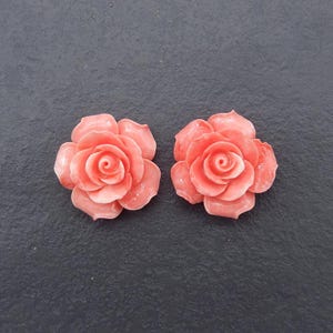 Puede incluir: Dos cabujones de flor de rosa de color coral rosa. Las flores están talladas en detalle y tienen un acabado liso y pulido.
