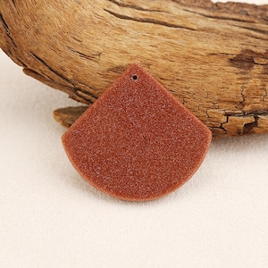 May include: A reddish-brown, fan-shaped pendant with a textured surface and a small hole at the top. The pendant is displayed against a backdrop of a piece of wood.