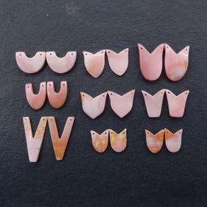May include: A collection of pink and peach-colored gemstone pendants in various shapes, including half-circles, shields, and tulip-like designs. Each pendant has a small hole for stringing. The pendants are arranged on a dark gray surface.