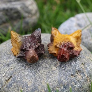 May include: Two carved stone wolf heads. One is primarily brown and yellow, the other is brown and purple. The wolf heads are resting on a grey stone, with green grass and other stones in the background. The wolf heads are detailed.
