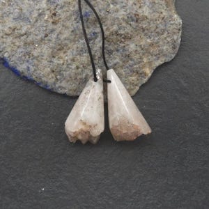 Puede incluir: Dos colgantes claros en forma de lágrima con una base rugosa y texturizada, suspendidos de un cordón negro. Los colgantes tienen una cualidad translúcida con toques marrones y están sobre un fondo gris texturizado.