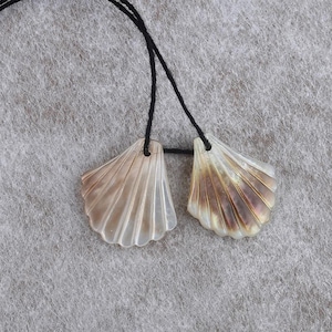 May include: Two shell pendants with iridescent colouring and a fan-like design, suspended from black cords. The shells have a pearlescent sheen, with shades of cream, tan, and brown. The pendants are displayed against a neutral grey background.