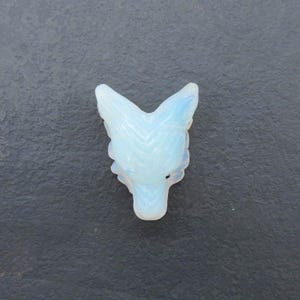 May include: A carved, opalescent wolf head pendant. The pendant is white with subtle blue hues, and the wolf's features are detailed. The pendant is set against a dark grey background.