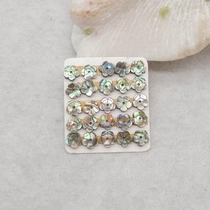 Puede incluir: Una colección de pequeñas cuentas iridiscentes en forma de flor, dispuestas sobre una tarjeta blanca. Las cuentas tienen un brillo nacarado con colores verdes, azules y plateados. Las cuentas son probablemente para la fabricación de joyas o proyectos de manualidades.