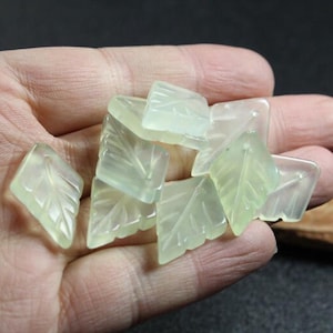 Puede incluir: Una colección de cuentas de piedra preciosa de color verde pálido, talladas en forma de hoja. Las cuentas son lisas y pulidas, con un delicado diseño de hoja.