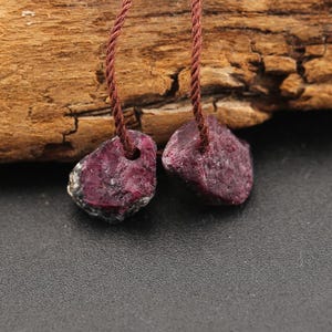 May include: Two raw, deep red ruby gemstones, each featuring a natural, irregular shape and a drilled hole at the top. Each stone is suspended by a thin, dark brown twisted cord, showcasing a rough, unpolished texture and dark crimson hues.