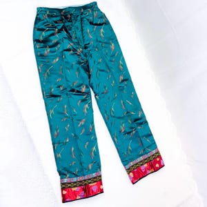 Peut inclure: Pantalon de pyjama en satin de couleur sarcelle avec un motif de plumes rouges, dorées et vertes. Les chevilles ont une bordure rouge et noire. Le tissu est brillant.