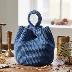 May include: A navy blue, handmade crochet bag with a rounded base and a looped handle. The bag has a textured, waffle-like stitch pattern and a drawstring closure. The bag is sitting on a wooden surface.