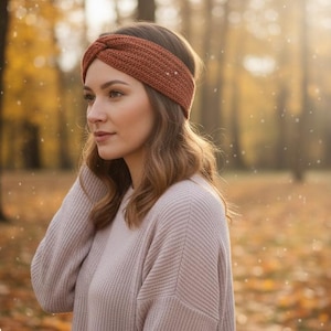 Könnte beinhalten: Ein rostfarbenes Stirnband mit Zopfmuster. Das Stirnband wird von einer Person mit langen, welligen, braunen Haaren getragen. Die Person trägt einen hellrosa Pullover. Der Hintergrund ist eine verschwommene Herbstszene mit Bäumen und fallenden Blättern.