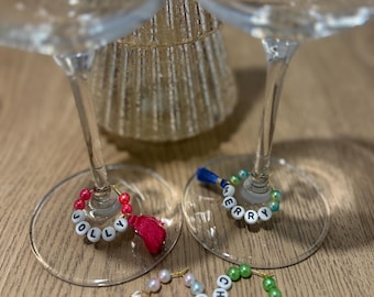 May include: Wine glass charms with white letter beads spelling "JOLLY", "MERRY", "HO HO HO", and "CHEERS". Each charm has a tassel in red, blue, white, and mint green. The charms are on the base of clear wine glasses.