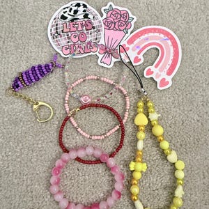 Könnte beinhalten: Eine Sammlung von Accessoires, darunter Armbänder, Aufkleber und ein Schlüsselanhänger. Die Armbänder sind in Rosa- und Rottönen gehalten und haben Perlen- und Steindesigns. Aufkleber zeigen eine Discokugel, einen Rosenstrauß und einen Regenbogen. Der Schlüsselanhänger ist lila.
