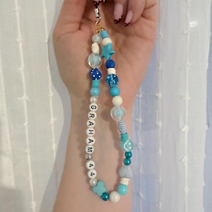 May include: A phone charm featuring a variety of blue and white beads, including a star-shaped bead, a blue die, a smiley face bead, and white letter beads spelling out GRAHAM followed by the number 44, all strung on a thin black cord.