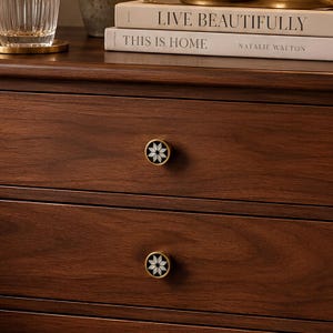 May include: A dark brown wooden dresser featuring two drawers with round brass-rimmed knobs. Each knob has a black circular inlay with a white star-shaped floral pattern. Two books are stacked on top, titled Live Beautifully and This Is Home.