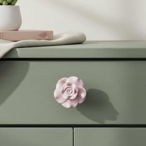 May include: A light pink, rose-shaped drawer pull is centered on a sage green drawer. The drawer pull is made of ceramic and has a detailed floral design. The drawer is part of a larger piece of furniture.