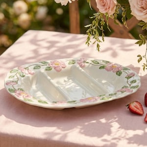 May include: An oval ceramic serving dish with a cream-colored glaze and a divided interior. The rim is decorated with a raised, three-dimensional relief pattern of pink wild roses and green leaves that wrap around the entire edge of the platter.