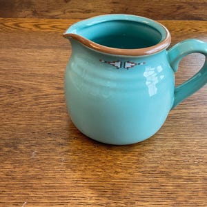May include: A turquoise ceramic pitcher with a rounded body and a handle. The rim is trimmed with a brown band and a decorative pattern. The pitcher is likely used for serving beverages.