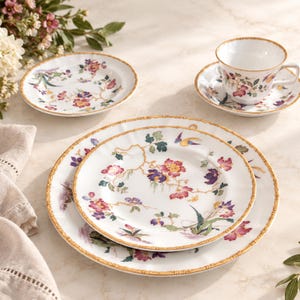 May include: A white porcelain dinnerware set featuring a delicate floral pattern in shades of pink, purple, and green with gold trim. The set includes a large dinner plate, a smaller salad plate, and a teacup with a matching saucer.