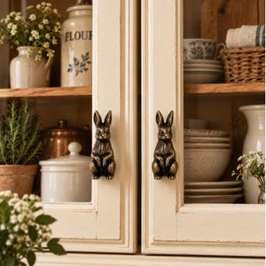 May include: Two antique brass-toned cabinet knobs shaped like upright rabbits. Each knob features a detailed, textured finish depicting a seated rabbit with pointed ears, serving as a functional handle on the glass-paneled cream cabinet doors.