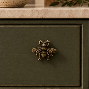 May include: A brass-toned metal cabinet knob shaped like a bee with detailed wings and body texture, mounted on a dark olive green drawer front. The bee figure features a segmented abdomen, six legs, and two antennae, providing a decorative handle.