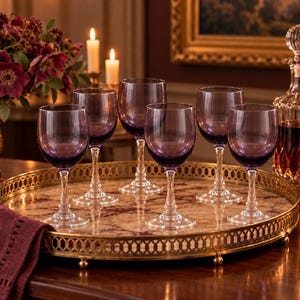 May include: A set of six purple glass wine glasses with clear stems and bases, arranged on a round gold metal tray with a decorative lattice rim and a marble-patterned surface. The glasses feature a deep violet hue and a classic rounded bowl.