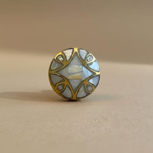 May include: A circular cabinet knob featuring a gold-toned metal frame with an intricate geometric pattern. The design is inlaid with iridescent white mother-of-pearl segments that fill the spaces between the metallic lines, creating a contrast.