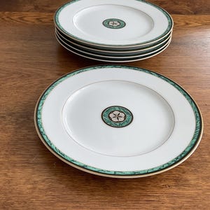 May include: A set of white ceramic plates with a green and gold border. Each plate features a central design of a green and white floral pattern. The plates are stacked and arranged on a wooden surface.