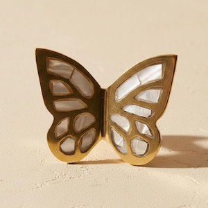 May include: A butterfly-shaped cabinet knob featuring a polished gold-tone metal frame with inlaid iridescent white mother-of-pearl segments filling the wing cutouts. The design displays a symmetrical wing pattern with a smooth metallic finish.