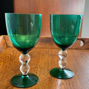 May include: Two emerald green wine glasses. Each glass features a rounded bowl, a clear, spherical stem, and a circular base. The glasses are made of glass and sit on a wooden surface. The glasses are empty.
