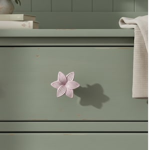 May include: A light pink, flower-shaped cabinet knob with six petals. The knob is mounted on a green painted wooden drawer. The drawer has horizontal lines and a distressed finish. A shadow of the knob is visible on the drawer.