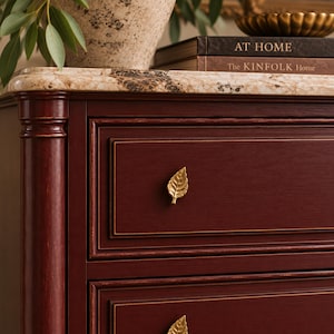 May include: A deep red wooden dresser featuring a marble top and gold-toned leaf-shaped drawer pulls. The dresser has two visible drawers with framed edges. Two books titled AT HOME and The KINFOLK Home are stacked on the marble surface.