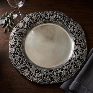 May include: Silver-colored decorative charger plate with an intricate grape vine and leaf design around the rim. The plate is round and sits on a dark wooden surface. A wine glass and napkin are also visible.