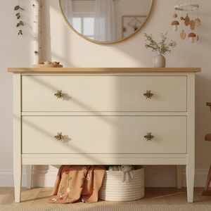 May include: A two-drawer cream-colored dresser with a light wood top and bee-shaped drawer pulls. A woven basket and a floral blanket are visible beneath the drawers. A round mirror and a mobile with wooden shapes are in the background.